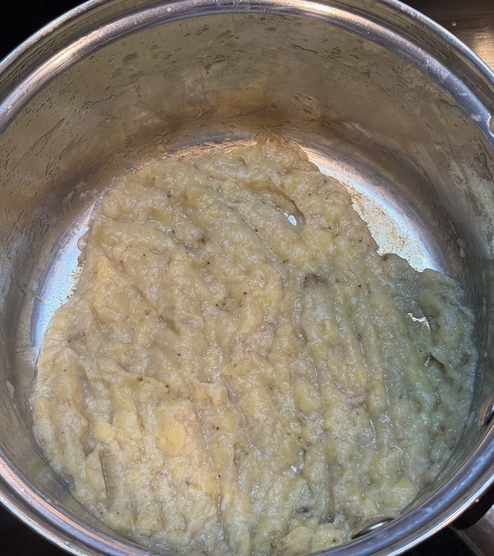 Pale yellow mashed banana paste spread in a deep stainless saucepan, holding spatula furrows