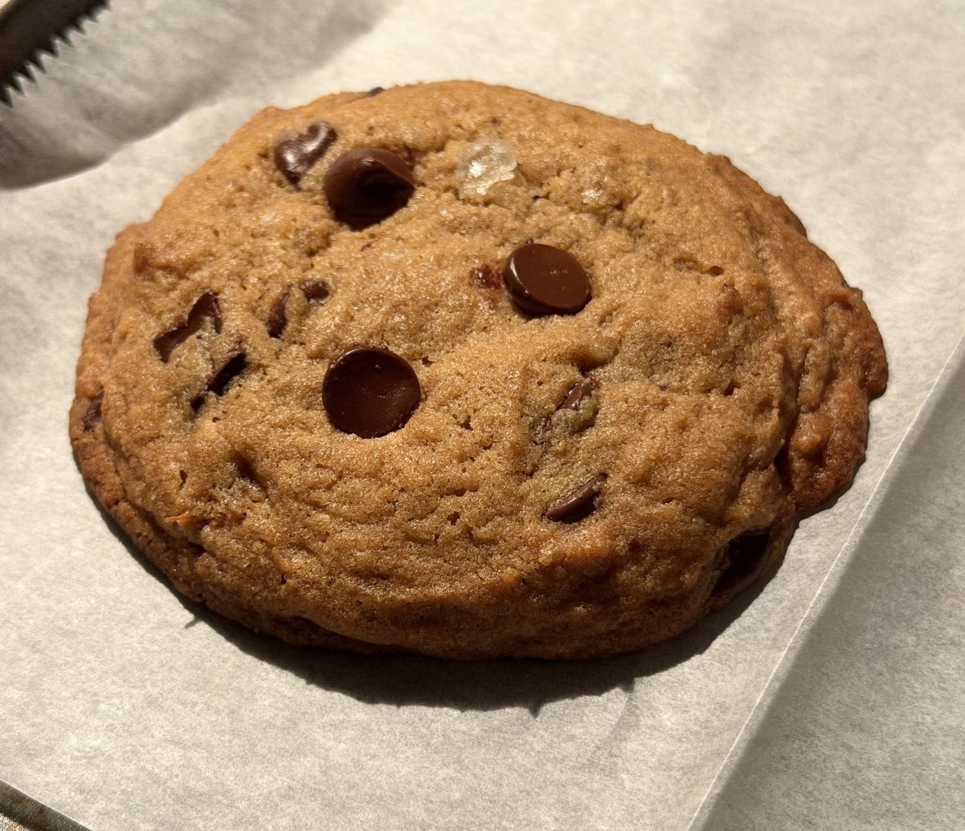A thick, domed brown butter banana cookie with dark chocolate chips and a flake of sea salt on parchment